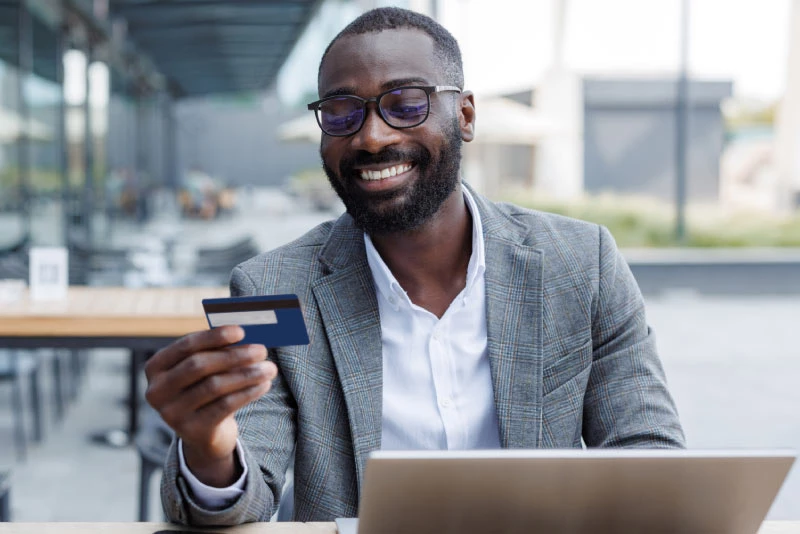 A photo of a man sitting on a patio with his laptop open as he smiles at his credit card